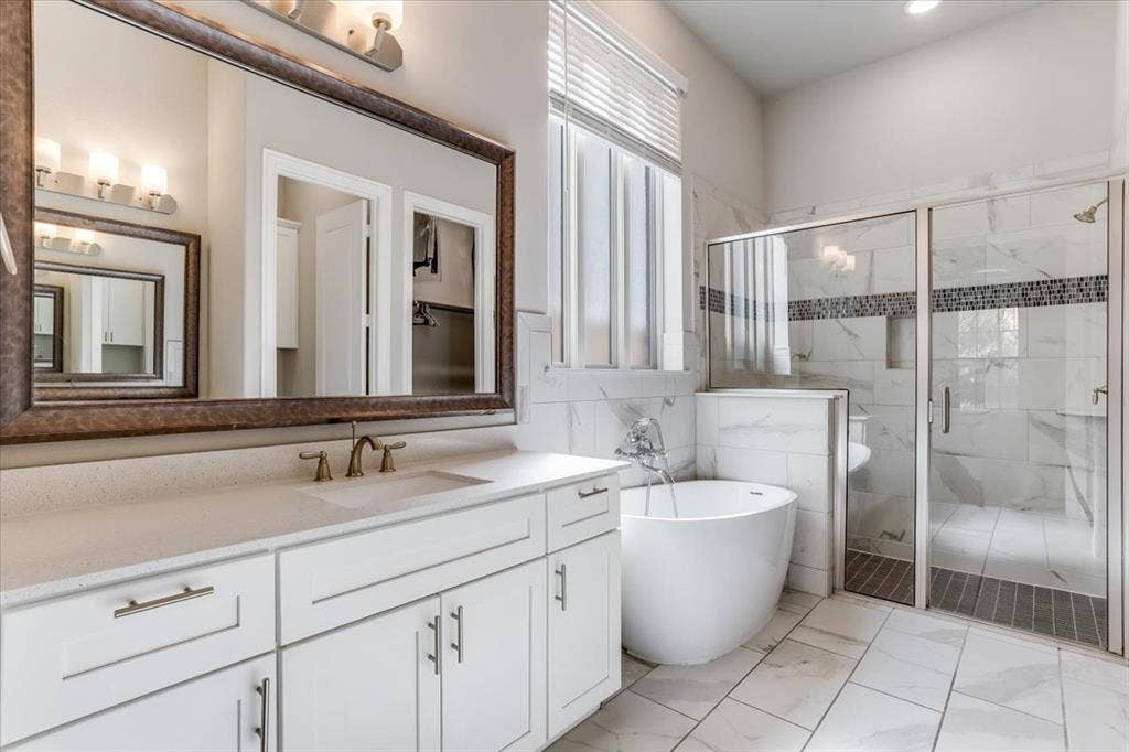Dual-sink vanity with white cabinetry and light-toned countertop, freestanding soaking tub, glass-enclosed shower with tiled walls, and large framed wall mirror
