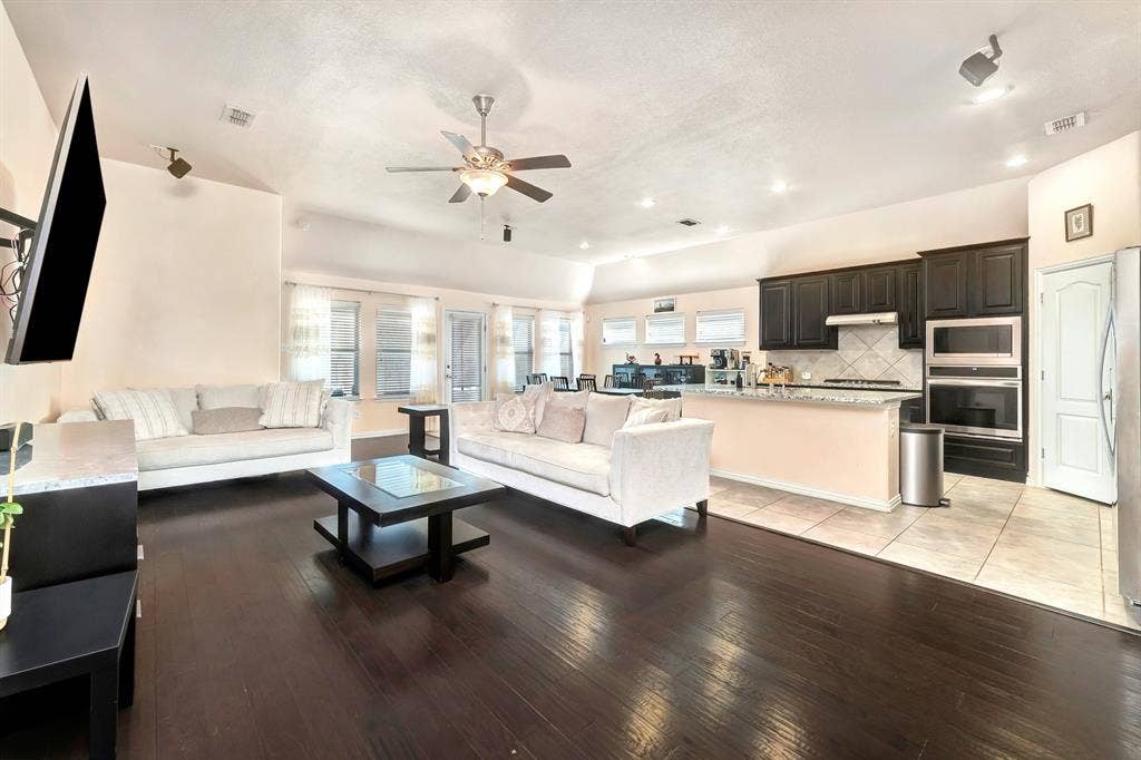 Open-concept area featuring dark wood-finish flooring, vaulted ceilings, a central ceiling fan, recessed lighting, and kitchen cabinetry with built-in stainless steel double ovens
