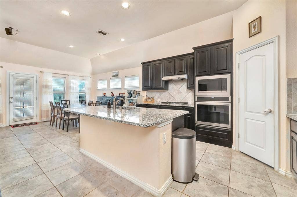 Kitchen featuring stone-finish countertops, dark wood cabinetry, integrated stainless steel double oven, tile backsplash, and ceramic tile flooring