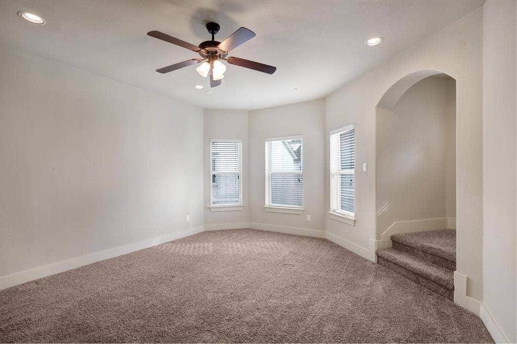 Carpeted room featuring a ceiling fan, recessed lighting, multiple windows with blinds, and an arched entryway leading to carpeted stairs