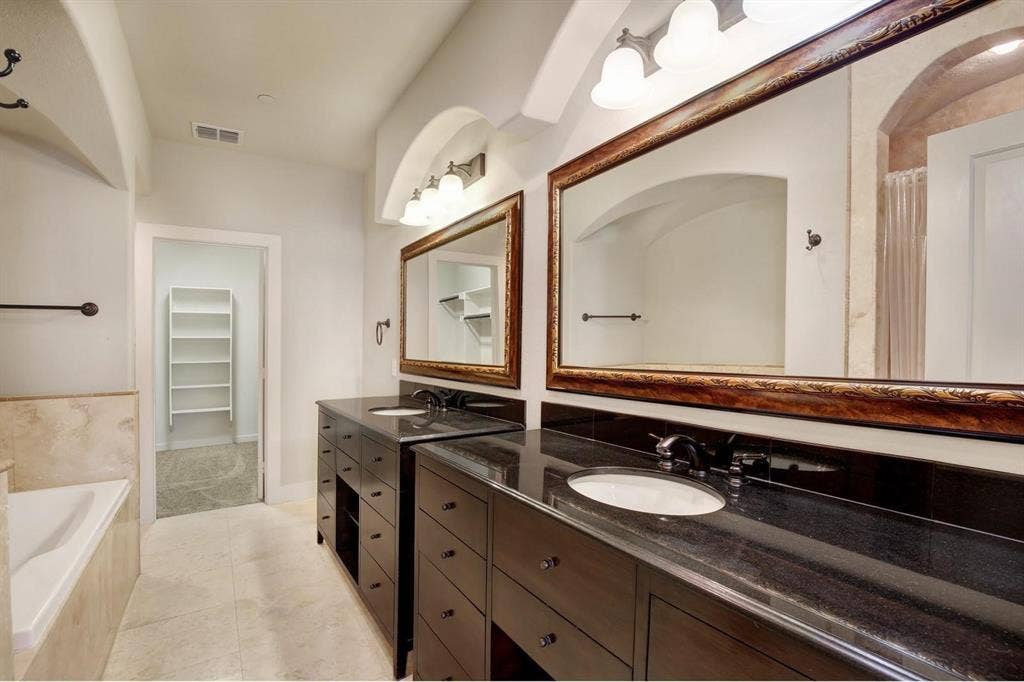 Dual dark wood vanities with undermount sinks and dark countertops, individual decorative framed mirrors, and a built-in bathtub with a tiled surround