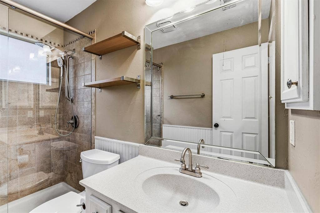 3916 Montrose Drive, Plano, TX 75025 Shower with glass enclosure, white vanity with integrated sink, large wall mirror, and two wood-finish wall shelves