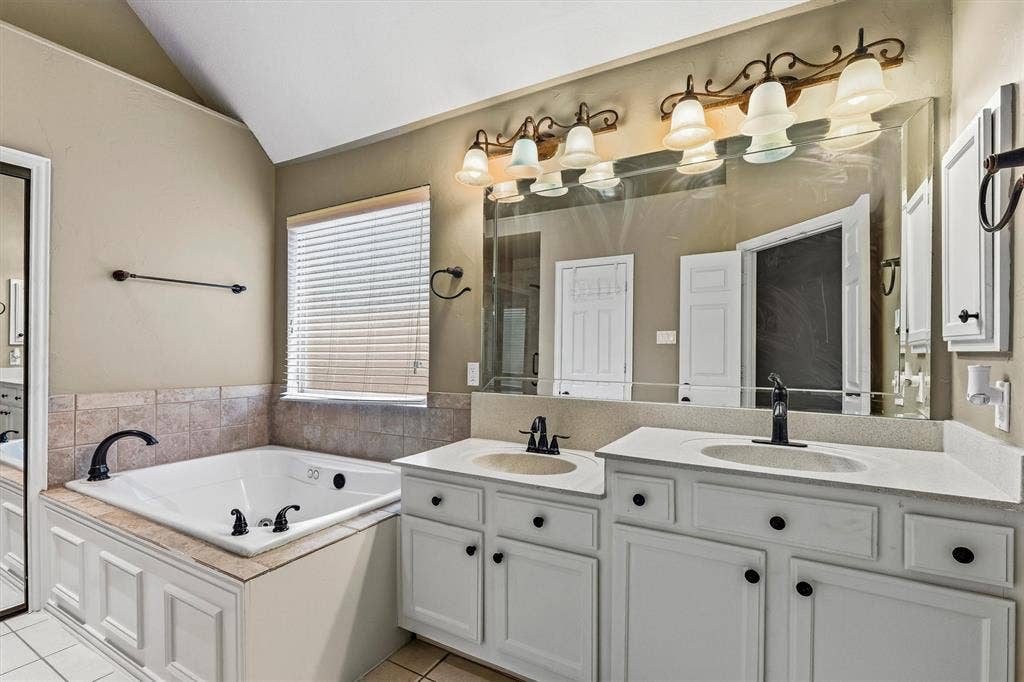 3916 Montrose Drive, Plano, TX 75025 Spa-inspired primary bathroom featuring a soaking tub with deck-mounted faucet and fixtures, dual-sink vanity with white cabinetry and brushed nickel hardware, quartz countertops, and a large framed mirror illuminated by a six-light vanity bar