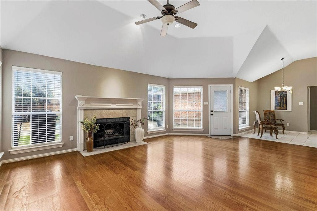 3916 Montrose Drive, Plano, TX 75025 Spacious great room showcasing gleaming hardwood flooring, a traditional fireplace with white mantel and surround, vaulted ceiling with ceiling fan, and abundant natural light from multiple windows and glass doors