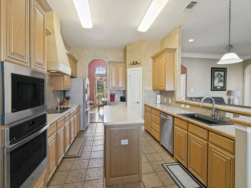 508 Singing Quail Trail, Haslet, TX 76052 Galley-style kitchen featuring wood cabinetry, tile flooring, stainless steel appliances including double wall ovens, a center island with wood-finish base, and granite countertops with undermount sink and chrome faucet