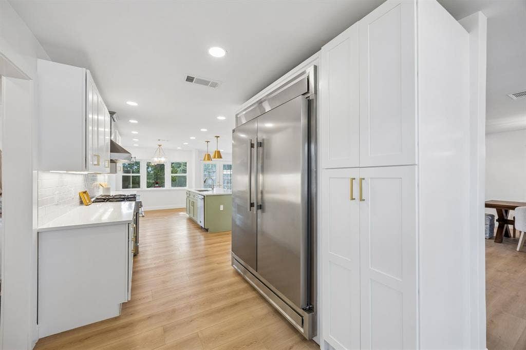11864 Cold Harbor Lane, Dallas, TX 75244 Chef's kitchen with a full size stainless refrigerator, crisp white cabinetry with gold pulls, and an open flow into the sunlit living spaces beyond.