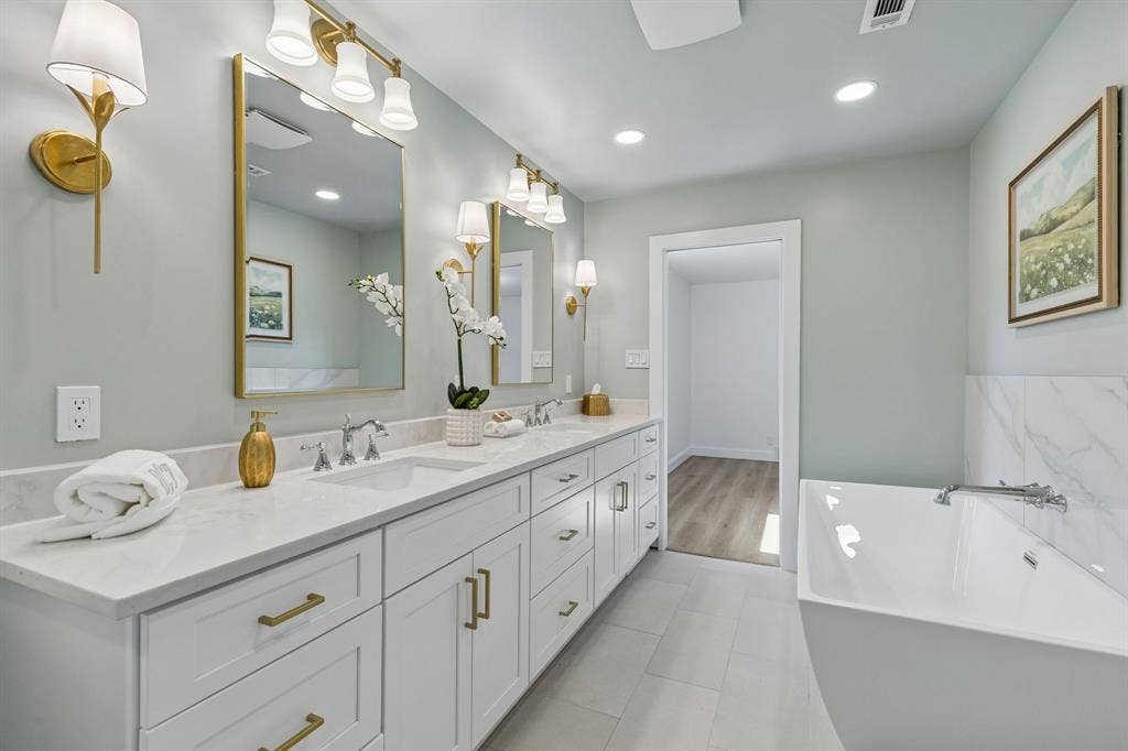 11864 Cold Harbor Lane, Dallas, TX 75244 Luxurious primary bath with dual vanities, brass framed mirrors, and elegant sconce lighting, plus a walk in closet just beyond.