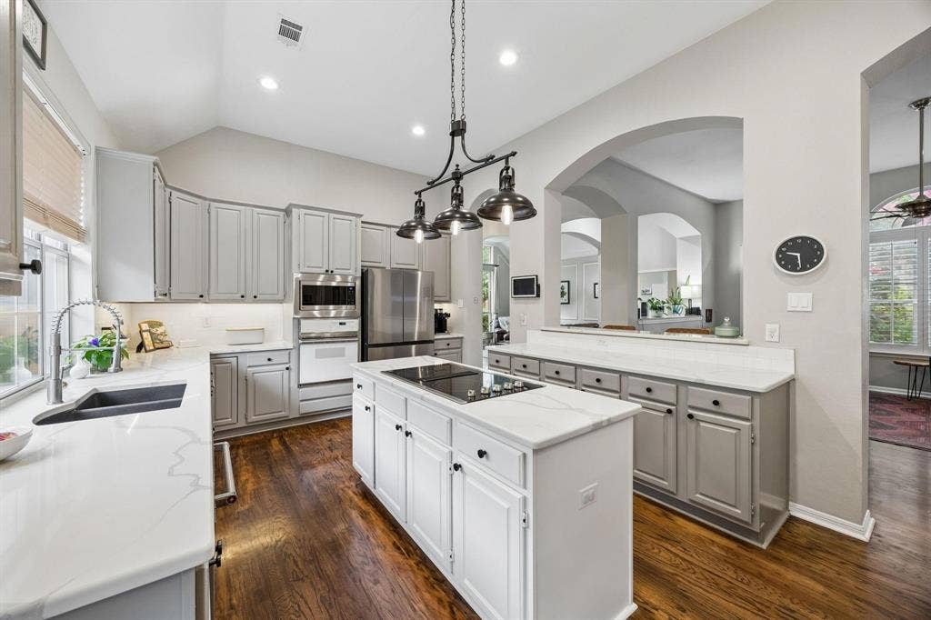 3104 Congress Avenue, Plano, TX 75025 Vaulted ceiling kitchen featuring an island cooktop, built-in stainless steel appliances, light-toned cabinetry, wood-finish flooring, and white countertops