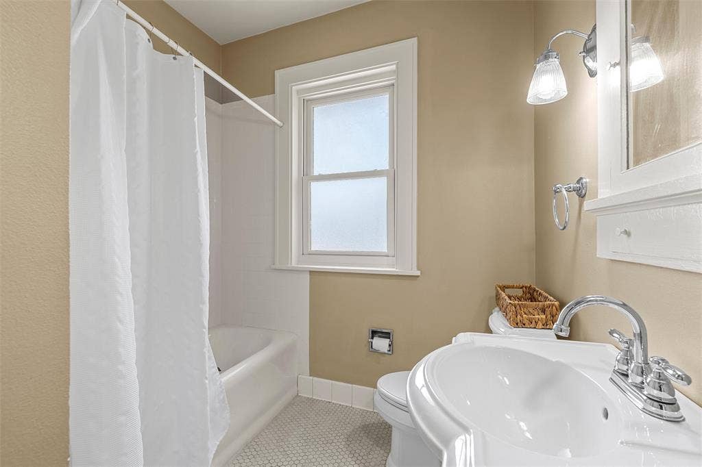 2424 Austin Avenue, Waco, TX 76701 Built-in bathtub with shower, pedestal sink with chrome faucet, window with white trim, wall-mounted vanity light, and hexagonal floor tiles