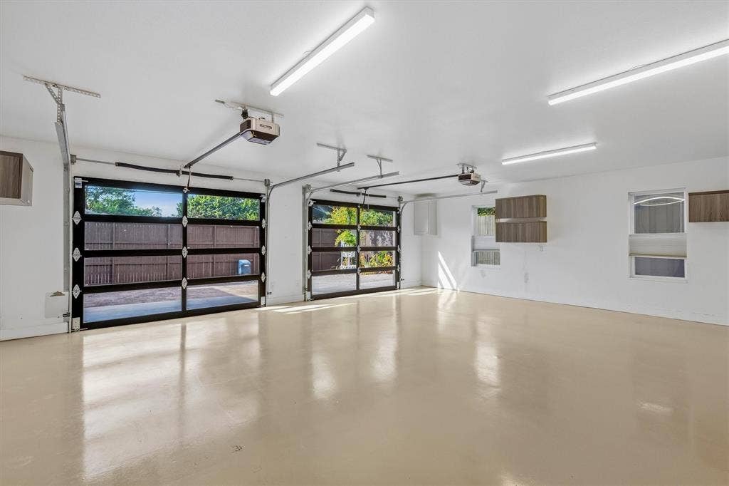 Two glass-paneled garage doors, two automatic garage door openers, multiple ceiling-mounted fluorescent light fixtures, two windows, and wall-mounted cabinetry