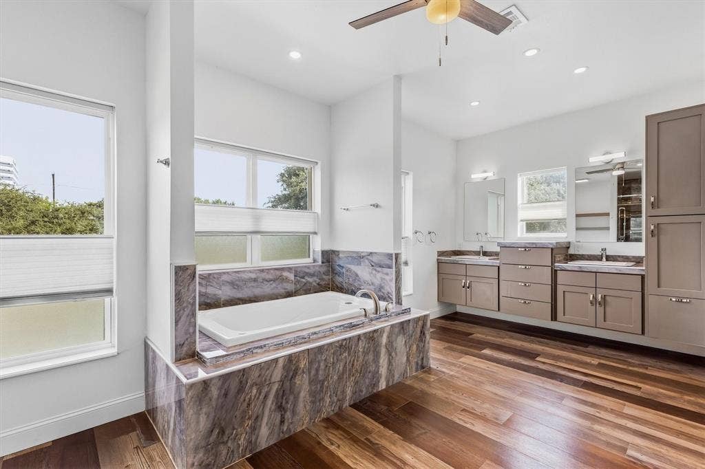 Spa-like bathroom with wood-finish flooring, a built-in tub with stone surround, dual vanities with integrated sinks, and abundant natural light from multiple windows