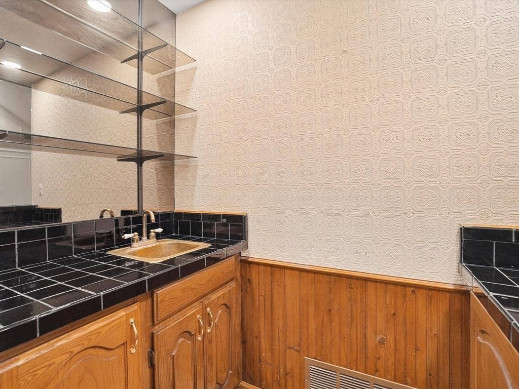 1304 Carriage Creek Drive, DeSoto, TX 75115 Built-in wet bar featuring a brass-tone sink, brushed nickel faucet, black tile countertop, wood cabinetry, and a mirrored backsplash with glass shelving