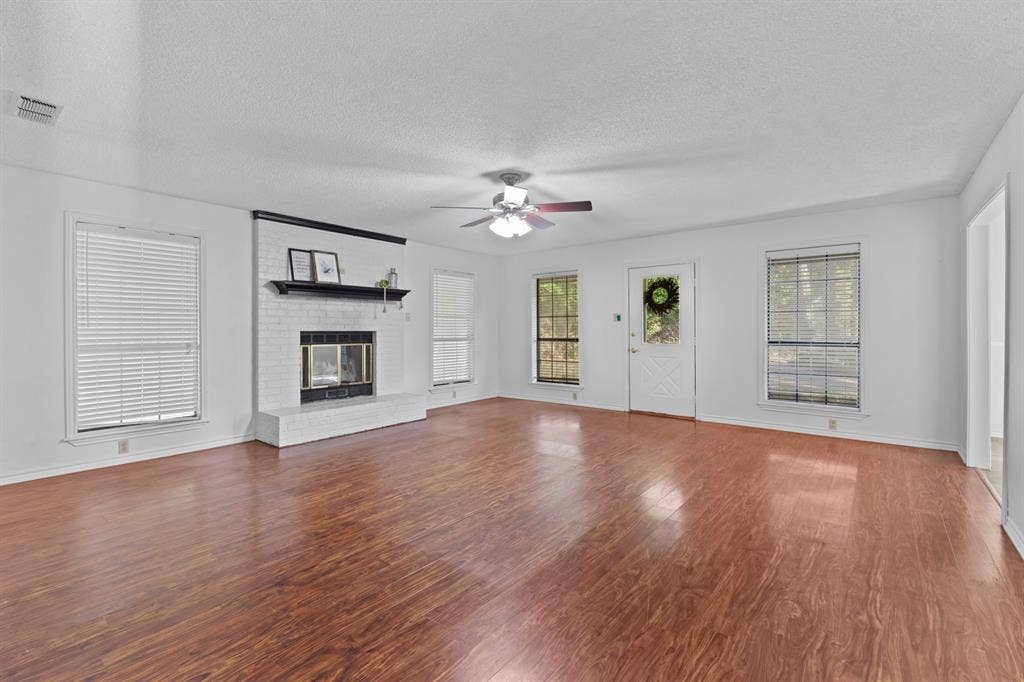 1656 Pineview Lane, Hideaway, TX 75771 Spacious room featuring wood-finish flooring, a brick-surround fireplace with a mantel, multiple windows, and a ceiling fan with integrated lighting