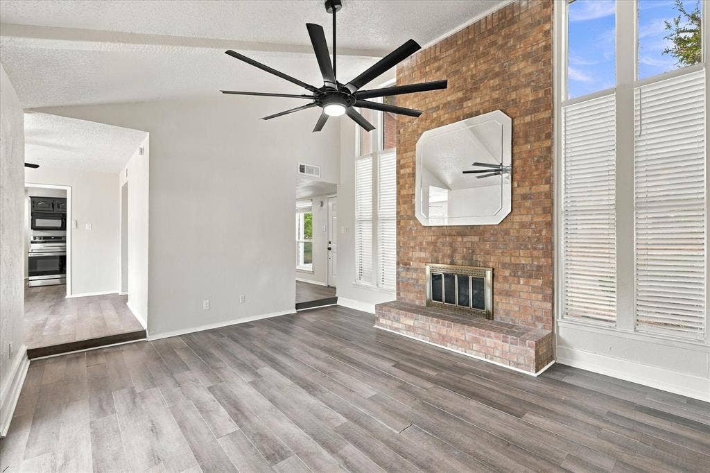 Vaulted ceiling featuring a contemporary ceiling fan, brick fireplace with a raised hearth, wood-finish flooring, and tall windows with interior shutters