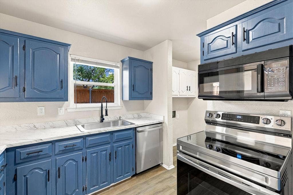 Kitchen featuring painted blue cabinetry, light-toned countertops, stainless steel sink with a matte black faucet, a stainless steel dishwasher, and a stainless steel range with an overhead microwave
