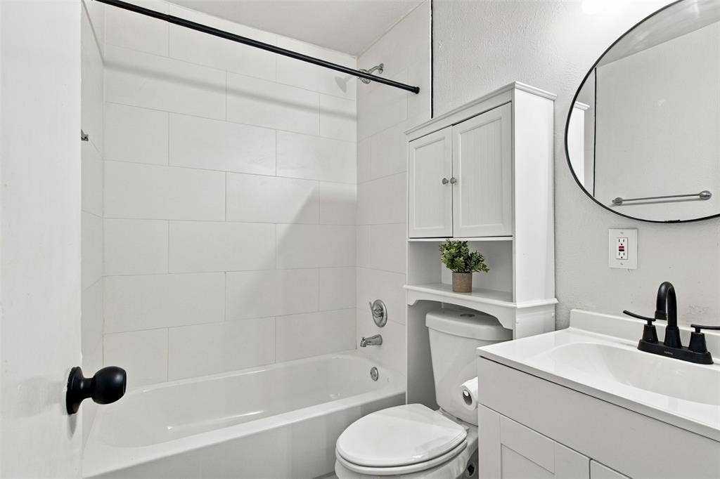 White tiled bathtub with a black shower rod, white vanity with an integrated sink, black two-handle faucet, circular mirror, and an over-the-toilet storage cabinet