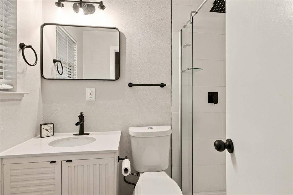 Corner vanity with a single basin and matte black faucet, glass-enclosed shower with black fixtures, white tile shower surround, and a rectangular framed mirror