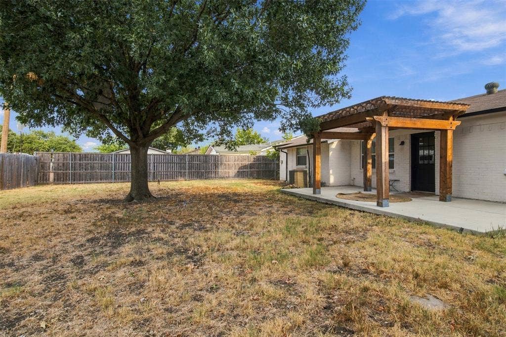 Expansive backyard featuring a mature tree, wood privacy fencing, concrete patio, and a wood pergola with a corrugated metal roof