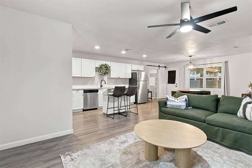 Open-concept living area featuring wood-finish flooring, white cabinetry, stainless steel appliances, and a modern ceiling fan
