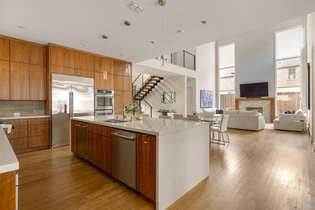 5724 Charlestown Drive, Dallas, TX 75230 Hardwood flooring throughout, a kitchen featuring wood cabinetry and stainless steel appliances, a large central island with seating, a living area with a stone fireplace, and a two-story ceiling