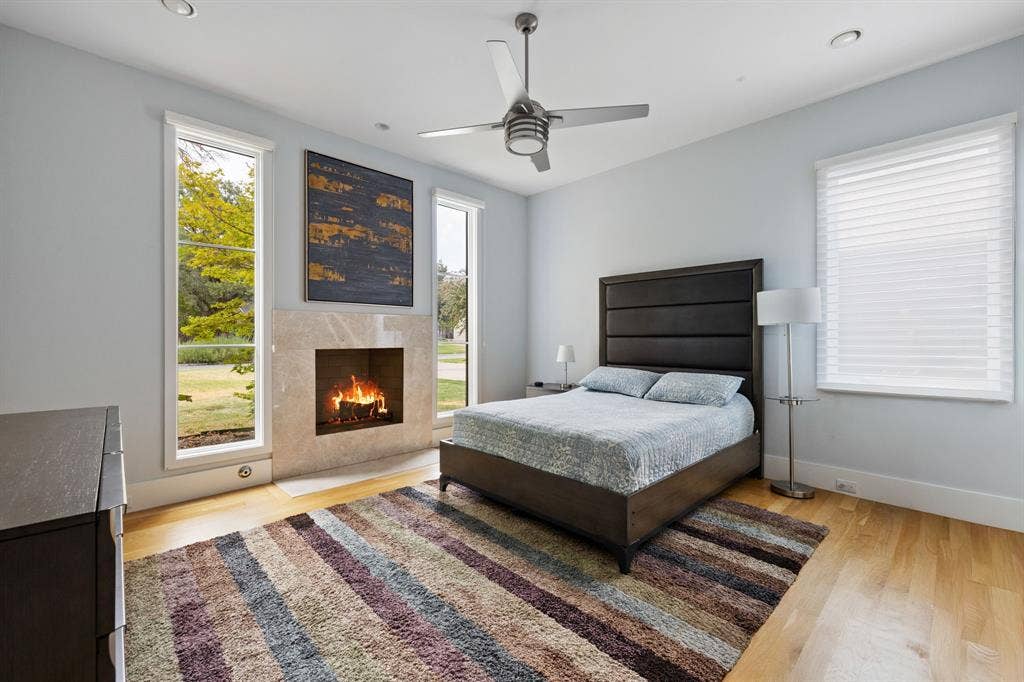 5724 Charlestown Drive, Dallas, TX 75230 Primary bedroom featuring a wood-finish floor, gas fireplace with stone surround, ceiling fan with integrated lighting, and abundant natural light from dual windows with motorized shades