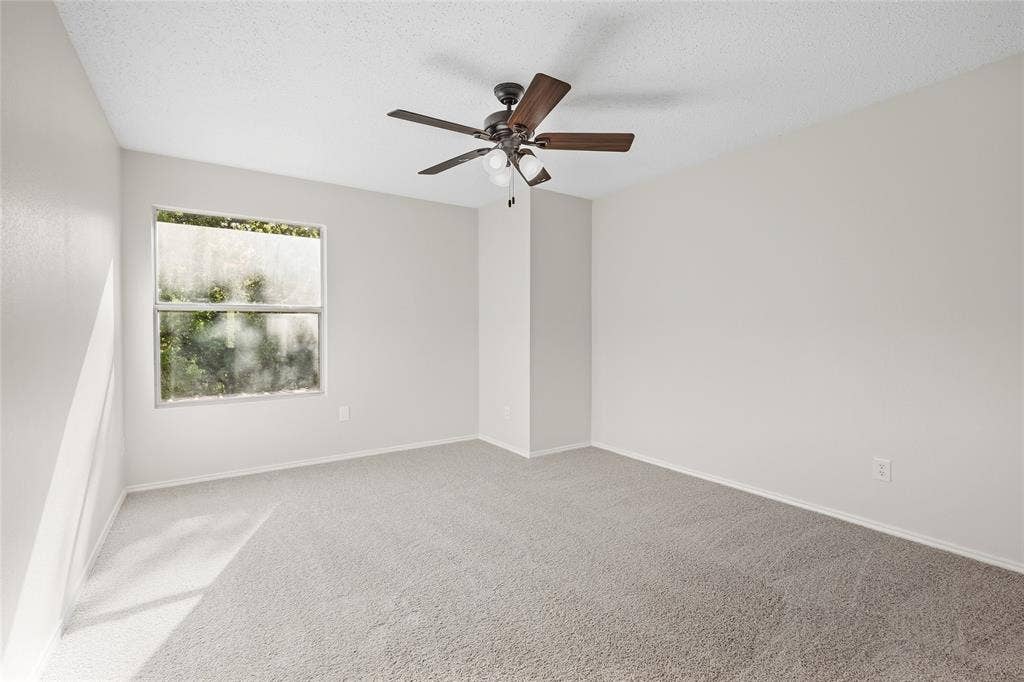 Spacious room featuring neutral carpeting, light-colored walls, a ceiling fan with integrated lighting, and natural light from a window