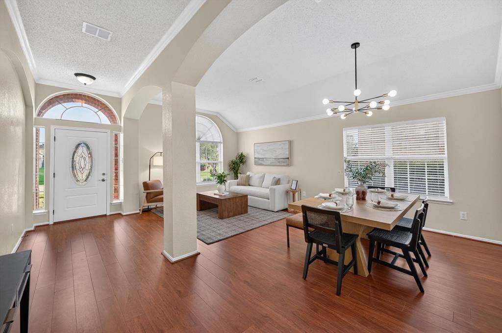 Open-concept living space featuring wood-finish flooring, vaulted ceilings, arched architectural entries, large decorative windows with brick surrounds, and a multi-bulb modern chandelier