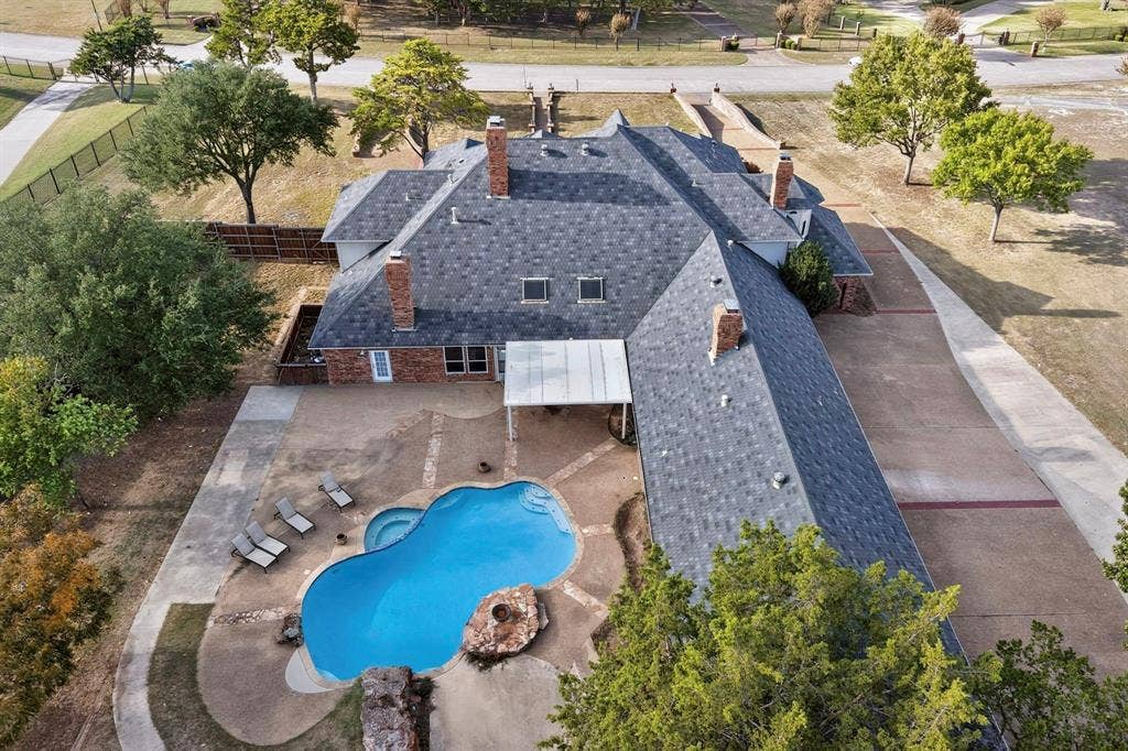 Aerial perspective featuring a multi-gabled brick residence with four chimneys, an expansive concrete patio, a swimming pool with integrated spa, and a covered outdoor terrace