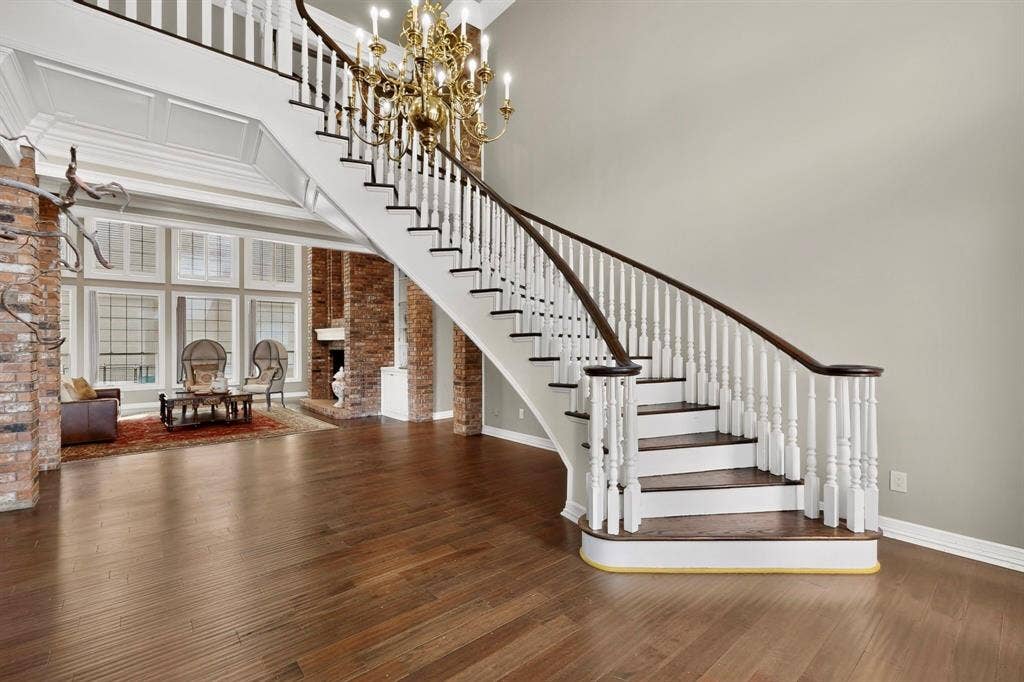 Curved staircase featuring white balusters, a dark wood handrail, and wood-finish treads, complemented by wide-plank wood-finish flooring, exposed brick structural pillars, and a multi-tiered metal chandelier