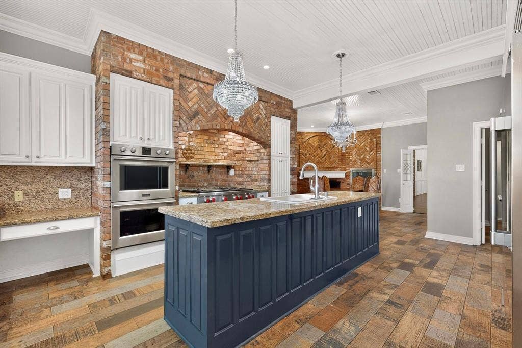 Kitchen featuring a blue-painted center island with a stone countertop, exposed brick wall with a built-in cooktop, stainless steel double wall ovens, beaded-board ceiling, and multi-toned wood-finish flooring