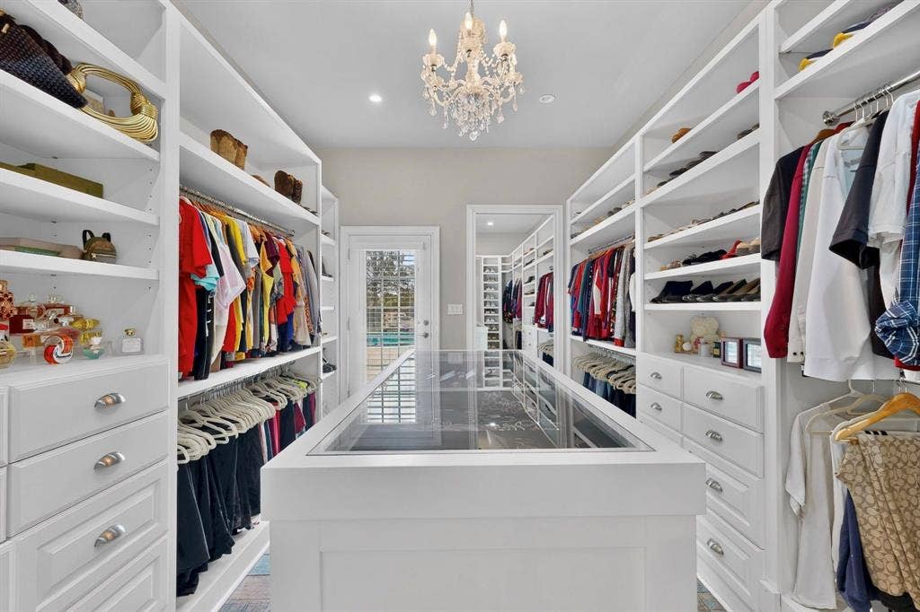 Custom walk-in closet featuring extensive white built-in shelving, a central island with a glass display top, recessed lighting, and a decorative chandelier