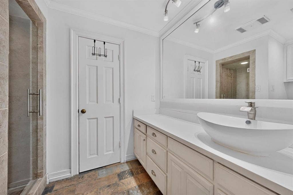 Bathroom featuring a vessel sink with a single-handle faucet, expansive wall-mounted mirror with integrated track lighting, shaker-style cabinetry with neutral countertop, and a walk-in shower with tile surround
