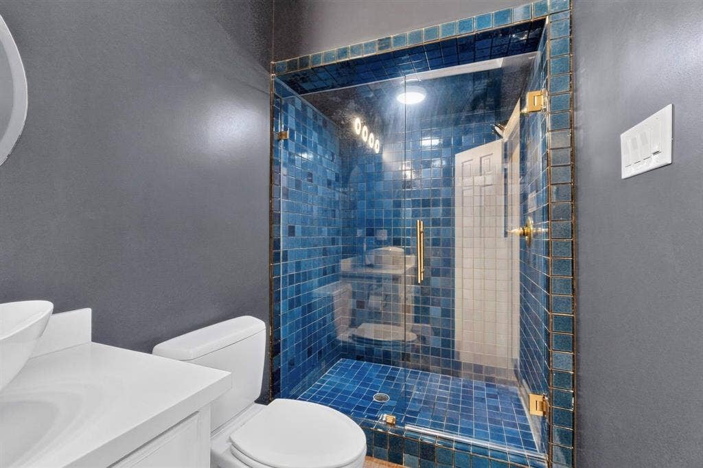 Walk-in shower featuring blue square mosaic tile, glass enclosure with brass-tone hardware, white vanity with integrated basin, and quartz-finish countertop