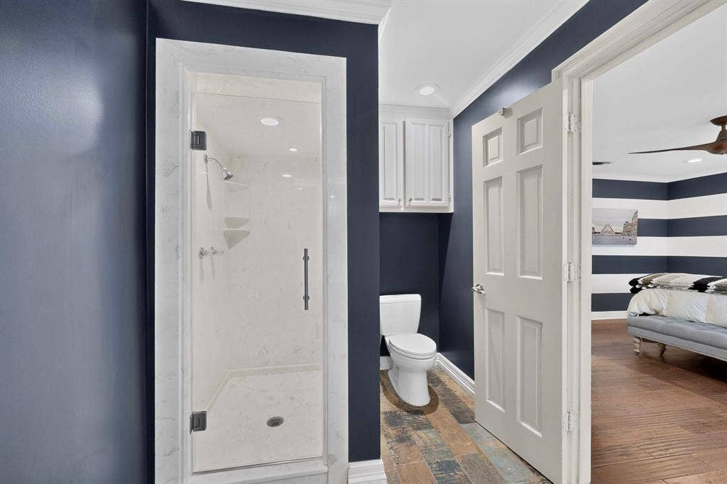 Shower enclosure with stone-patterned surround, white wall-mounted cabinetry, floor-to-ceiling dark-toned wall finish, and wood-finish flooring