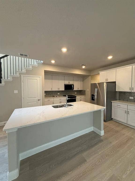 Kitchen featuring a center island with sink, stainless steel appliances, upper and lower cabinetry, and wood-finish flooring