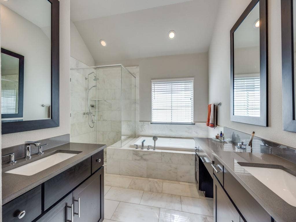 Dual vanities with integrated sinks and dark cabinetry, large soaking tub, glass-enclosed shower with tiled walls, natural light from a privacy window, and recessed lighting