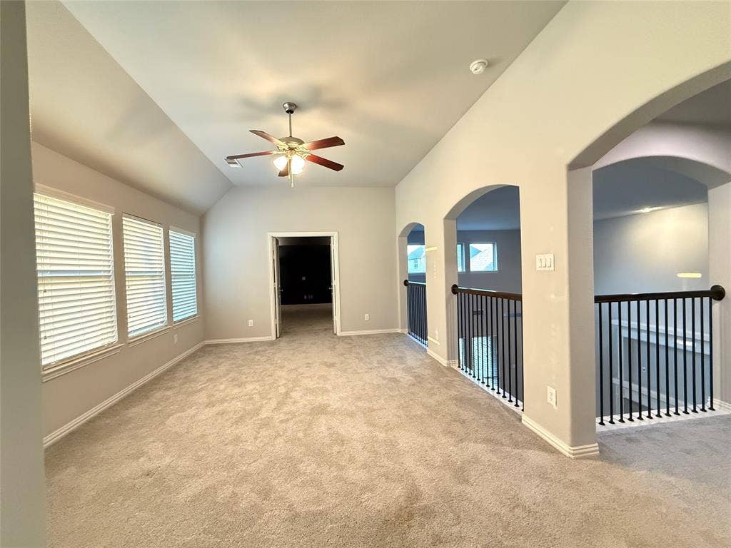 11818 Antler Drive, Frisco, TX 75035 Carpeted room featuring a vaulted ceiling with a ceiling fan, multiple windows with blinds, a doorway, and arched openings to a balustrade with black spindles