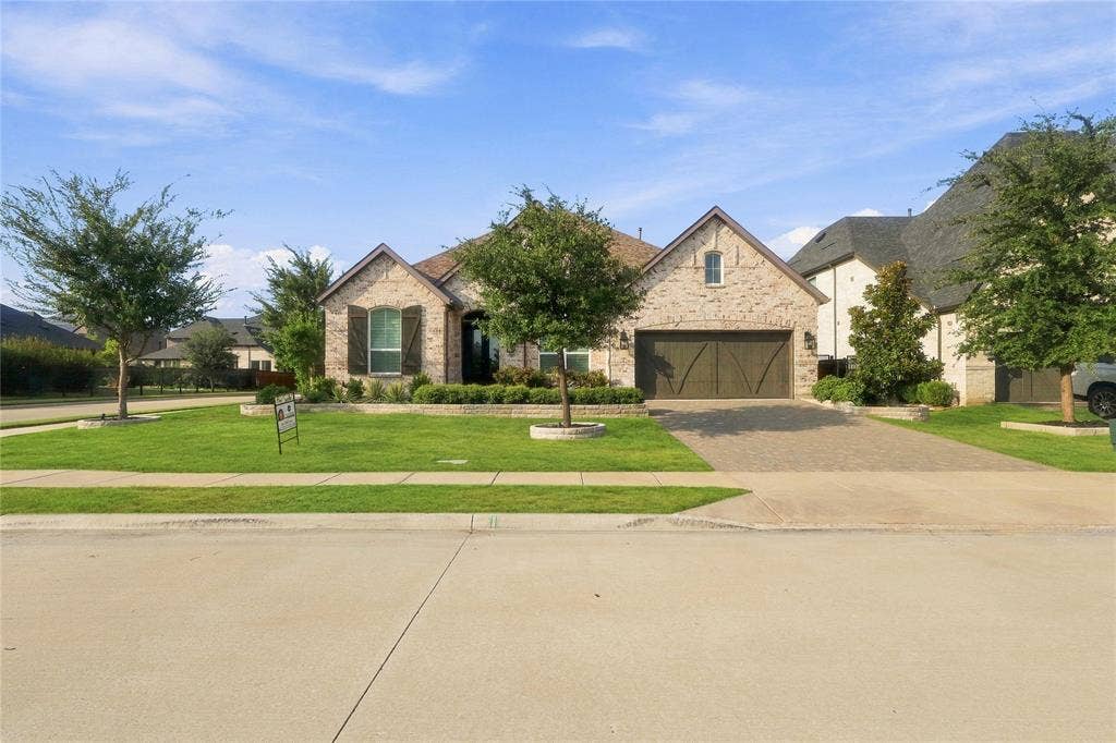 Stone landscape border with dark wood shutters, attached garage with wood-finish door, paver driveway, and front lawn with mature trees
