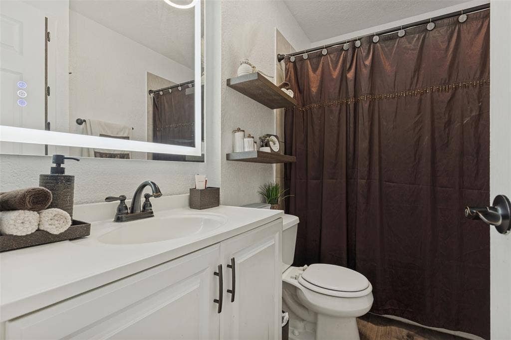 1522 Carroll Drive, Garland, TX 75041 White vanity with a solid-surface countertop, under-mount sink, matte black hardware, integrated LED-backlit mirror, and tiered wood-finish floating shelving