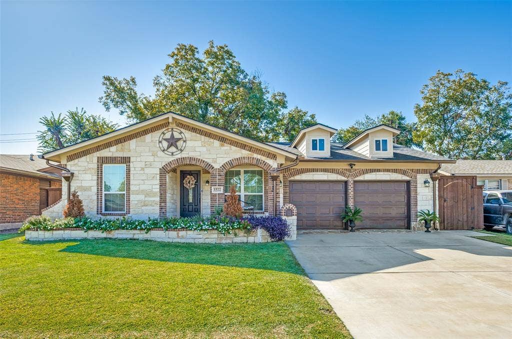 1522 Carroll Drive, Garland, TX 75041 Single-story stone and brick exterior featuring dual arched garage bays, dormer windows, and an elevated stone garden bed