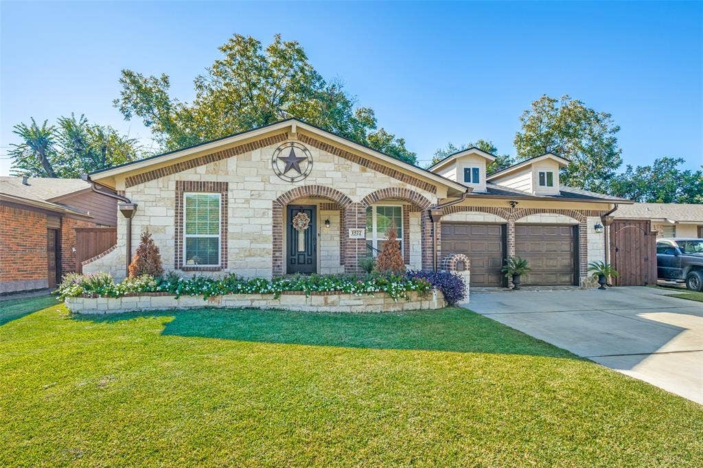 1522 Carroll Drive, Garland, TX 75041 Stone-clad exterior featuring arched brick entryways, two-car garage with wood-finish doors, and a raised stone masonry garden bed