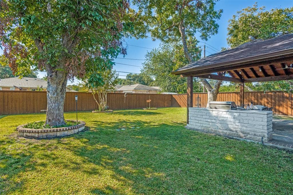 1522 Carroll Drive, Garland, TX 75041 Backyard featuring a covered patio with a built-in masonry grill station, a brick-enclosed tree planter, and perimeter wood fencing