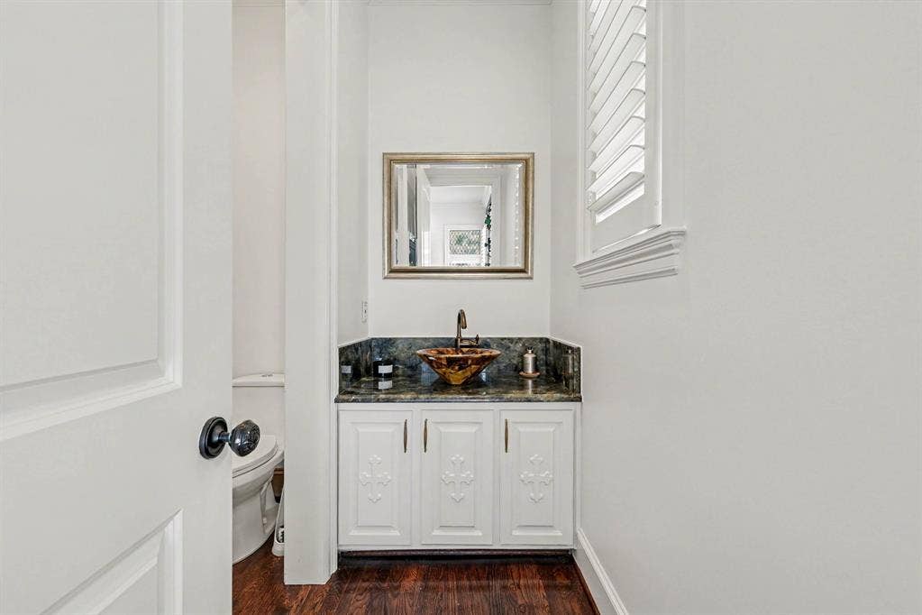 5542 Monticello Avenue, Dallas, TX 75206 Powder room featuring a decorative vessel sink, dark stone countertop, white cabinetry with decorative panels, wood-finish flooring, and window with plantation shutters