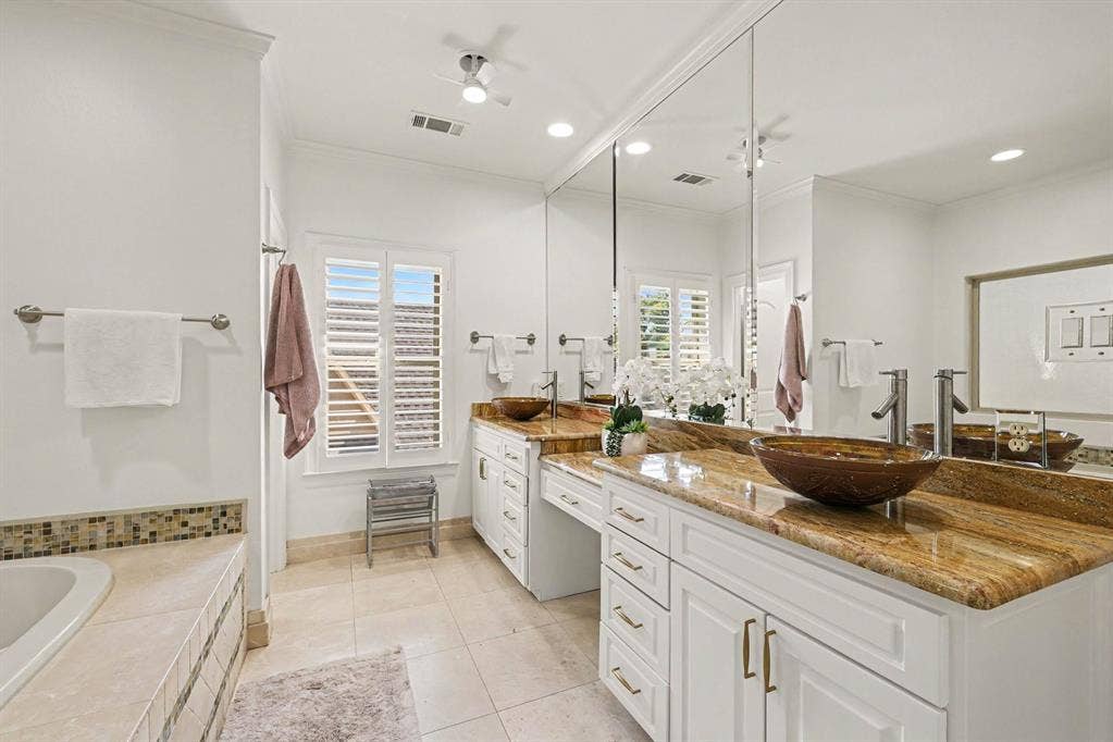 5542 Monticello Avenue, Dallas, TX 75206 Dual vessel sinks set on a stone countertop with white cabinetry, extensive wall-mounted mirrors, and ceiling fan