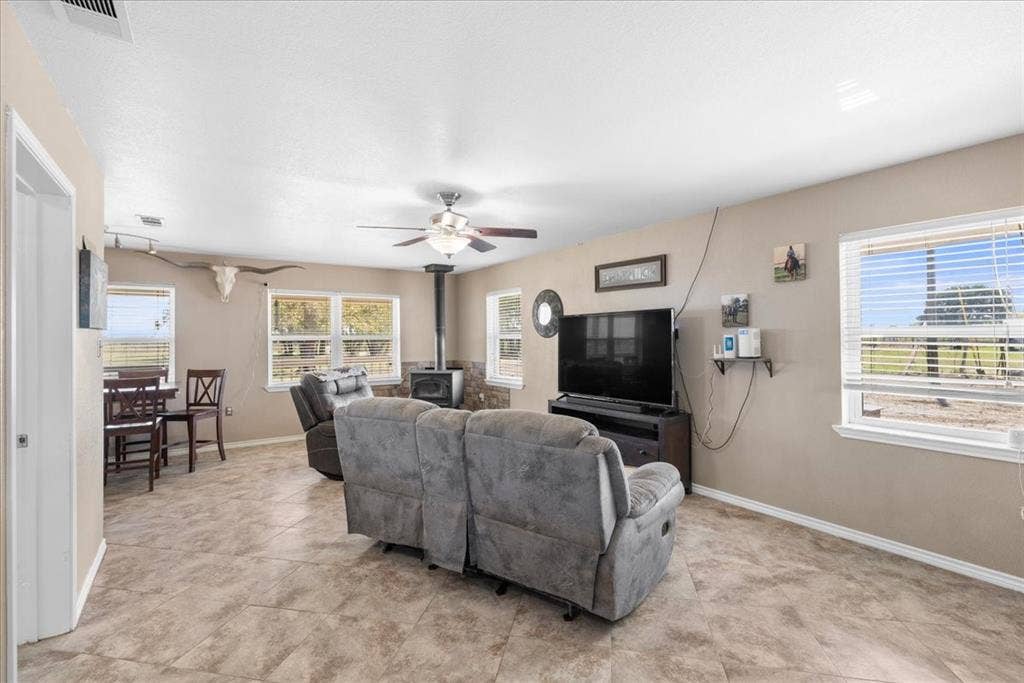 Open-concept interior featuring a wood-burning stove with stone-finish surround, multiple windows with blinds, a ceiling fan, and tile flooring