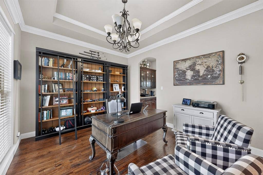 5020 Lindy Court, Midlothian, TX 76065 Sophisticated home office featuring rich wood-finish flooring, extensive built-in cabinetry with glass-front display shelving, a traditional desk with decorative turned legs, coffered ceiling with crown molding, and a coordinating upholstered accent chair