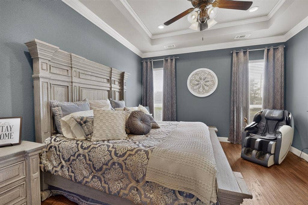 5020 Lindy Court, Midlothian, TX 76065 Master bedroom featuring soft blue walls, a decorative tray ceiling with ceiling fan, hardwood flooring, and large windows with coordinating drapery