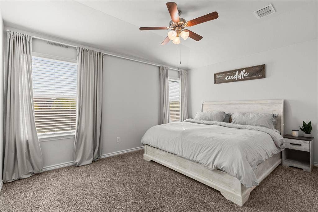 Neutral wall finishes, carpeted flooring, two windows with blinds, and a ceiling fan with integrated lighting (primary bedroom actual)