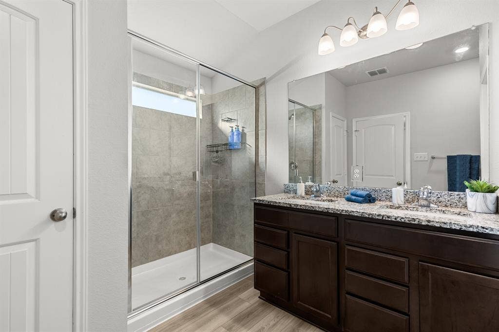 Spacious bathroom featuring a dual vanity with a patterned countertop, dark wood-finish cabinetry, a large frameless mirror, and a walk-in shower with tile surround