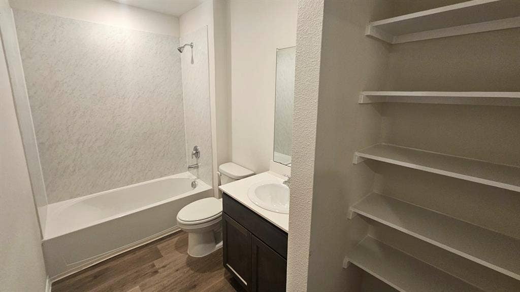 Bathroom featuring a full tub and shower combination with a textured surround, a single vanity with a dark wood-finish cabinet and white countertop, a white toilet, and wood-finish flooring